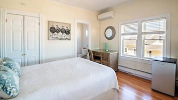 A bedroom with a bed, a desk, a refrigerator, a window with blinds, and a door.