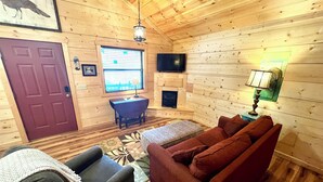 A cozy cabin interior with a wooden floor, a sofa, a chair, a TV, a fireplace, and a window with blinds. - Turkey Trot #73 Cabin in town, walking distance to courthouse square. (Mountain View)