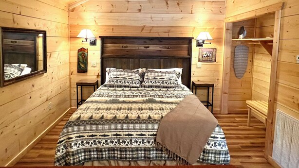A wooden cabin bedroom with a bed, two bedside lamps, a headboard, a mirror, and a wall-mounted shelf. - Turkey Trot #73 Cabin in town, walking distance to courthouse square. (Mountain View)
