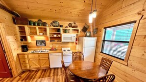 A wooden cabin kitchen with a dining table, chairs, a refrigerator, a stove, and shelves with various kitchen items. - Turkey Trot #73 Cabin in town, walking distance to courthouse square. (Mountain View)