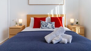 Un lit bien fait, recouvert d’une couverture bleue, avec des oreillers rouges et blancs, et une serviette blanche posée sur le côté. On y trouve deux tables de chevet avec des lampes et de petites plantes.
