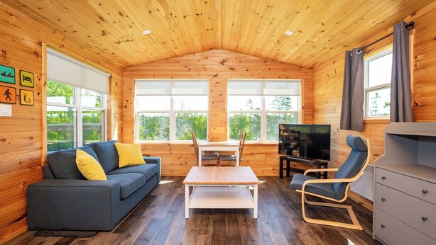 A cozy cabin interior with a wooden ceiling, a sofa with cushions, a dining table, a TV, and a dresser.