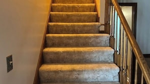 Un escalier recouvert de moquette, avec des rampes en bois et un interrupteur sur le mur.