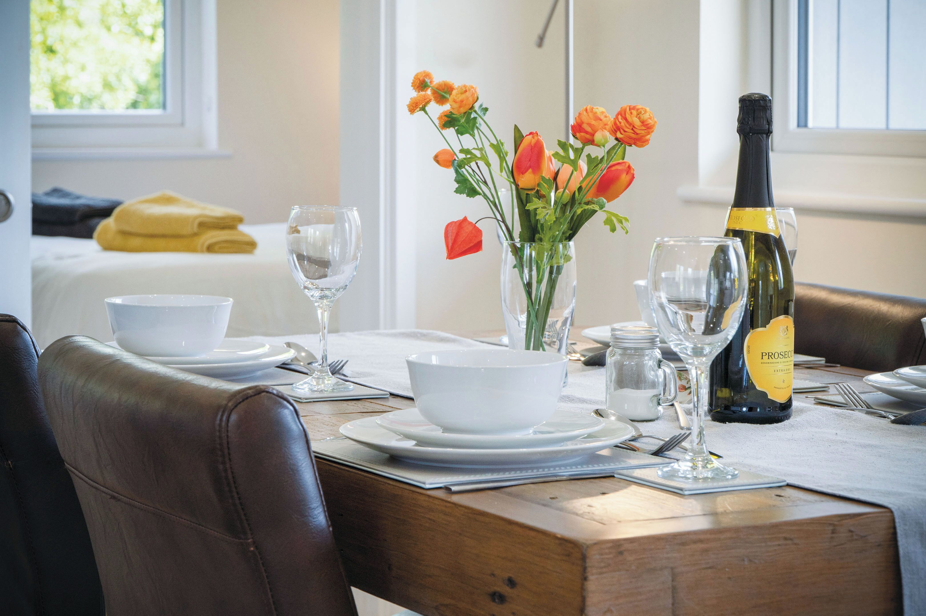 Une table à manger dressée avec des bols blancs, des verres à vin, une bouteille de Prosecco et un vase rempli de fleurs oranges.