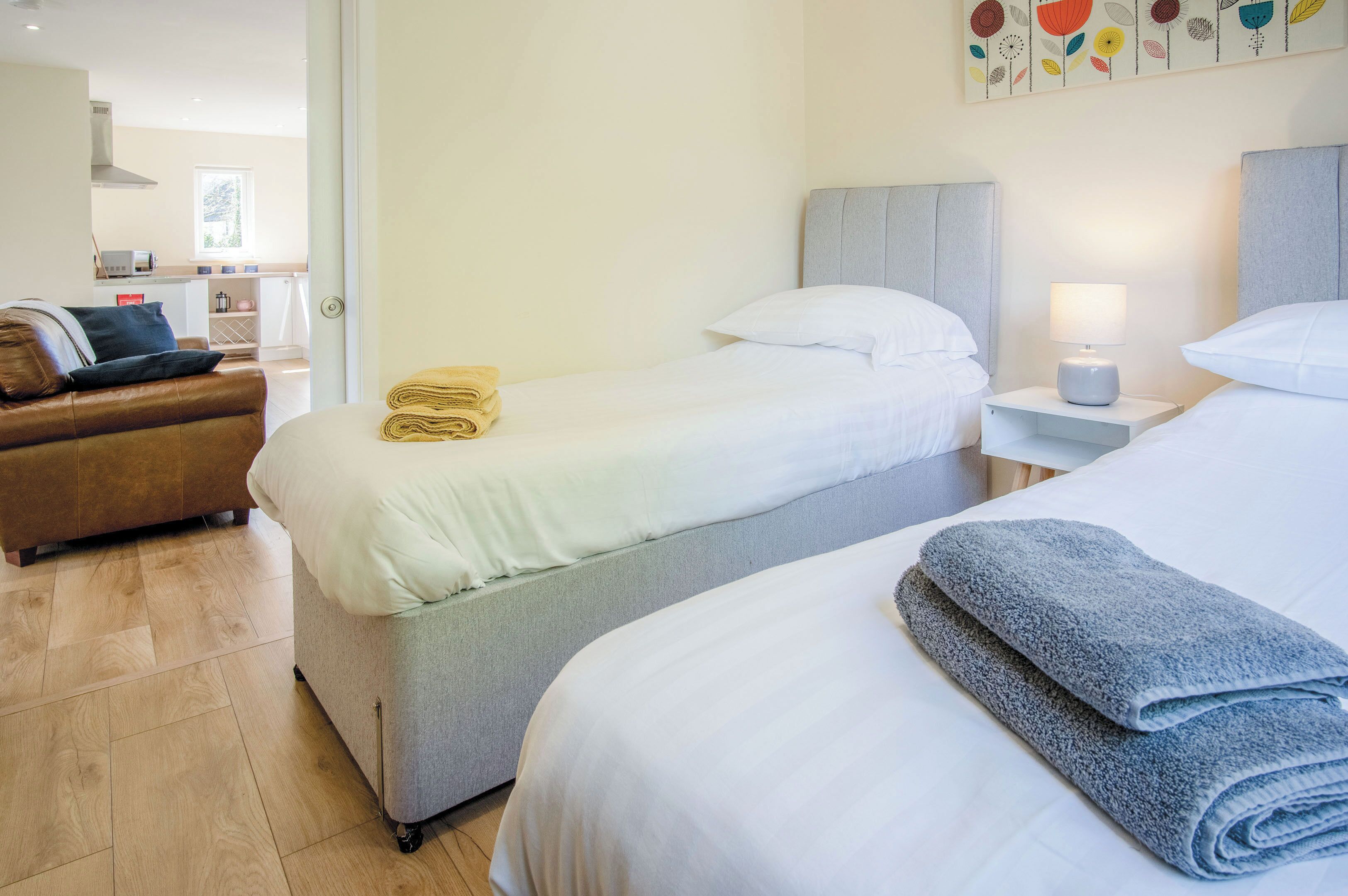 A hotel room with two beds, a sofa, a small table with a lamp, and a kitchen in the background.
