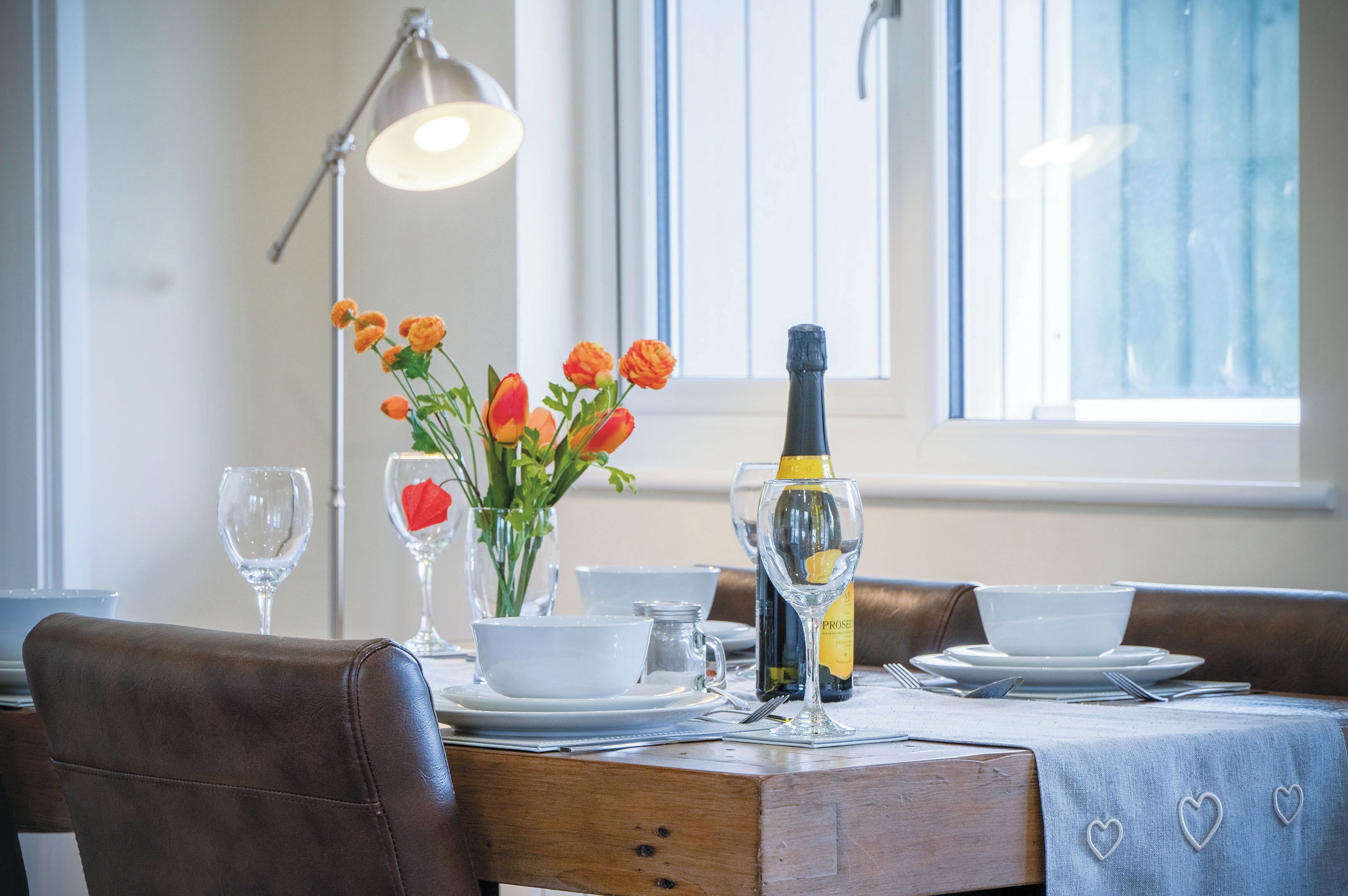 Una mesa de comedor dispuesta con platos blancos, tazones y una botella de champán. Hay flores naranjas en un florero, y se ve una lámpara al fondo.