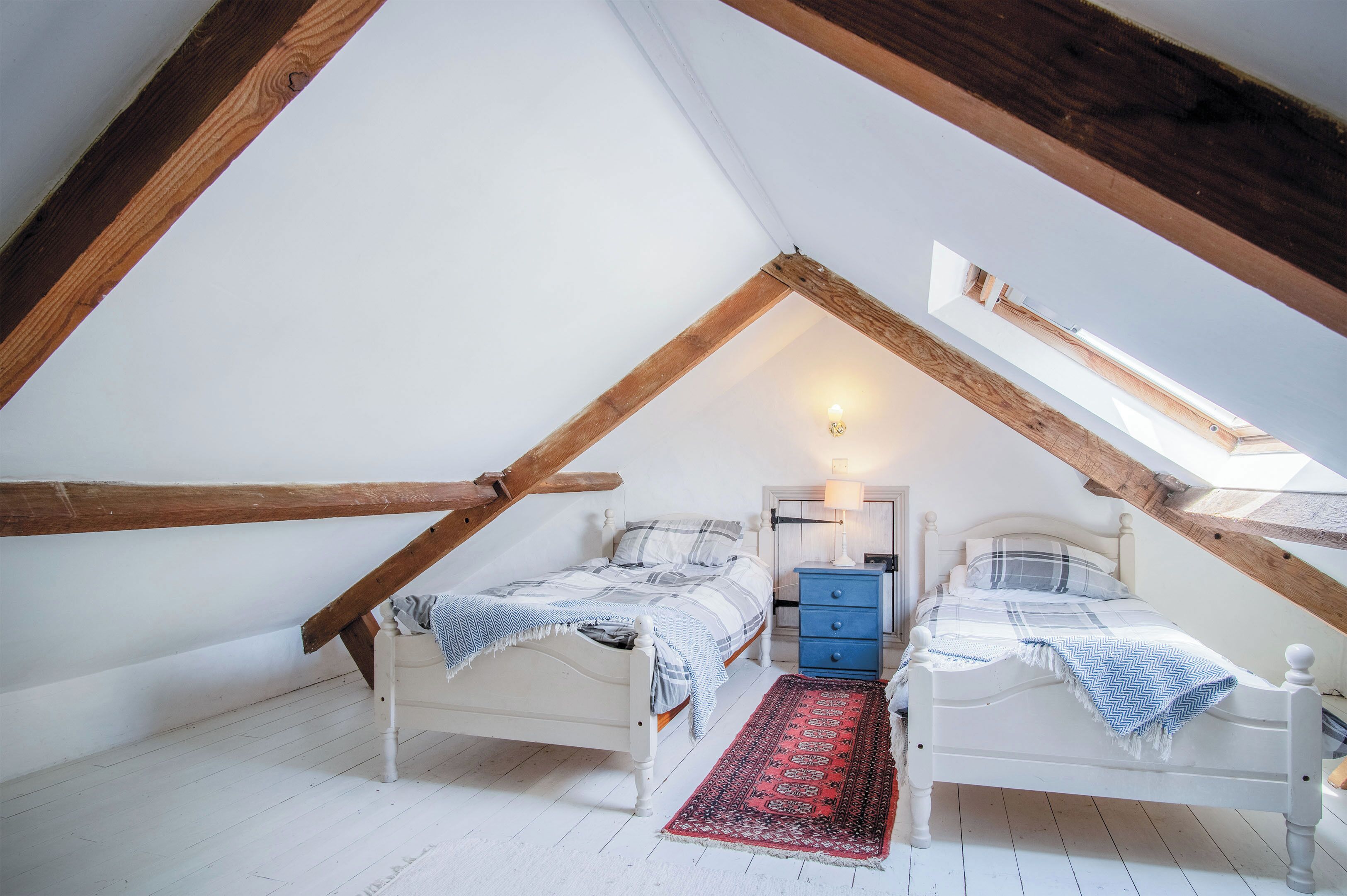 Une petite chambre mansardée avec deux lits, une table de chevet bleue, un tapis à motifs rouges et noirs et des poutres en bois.