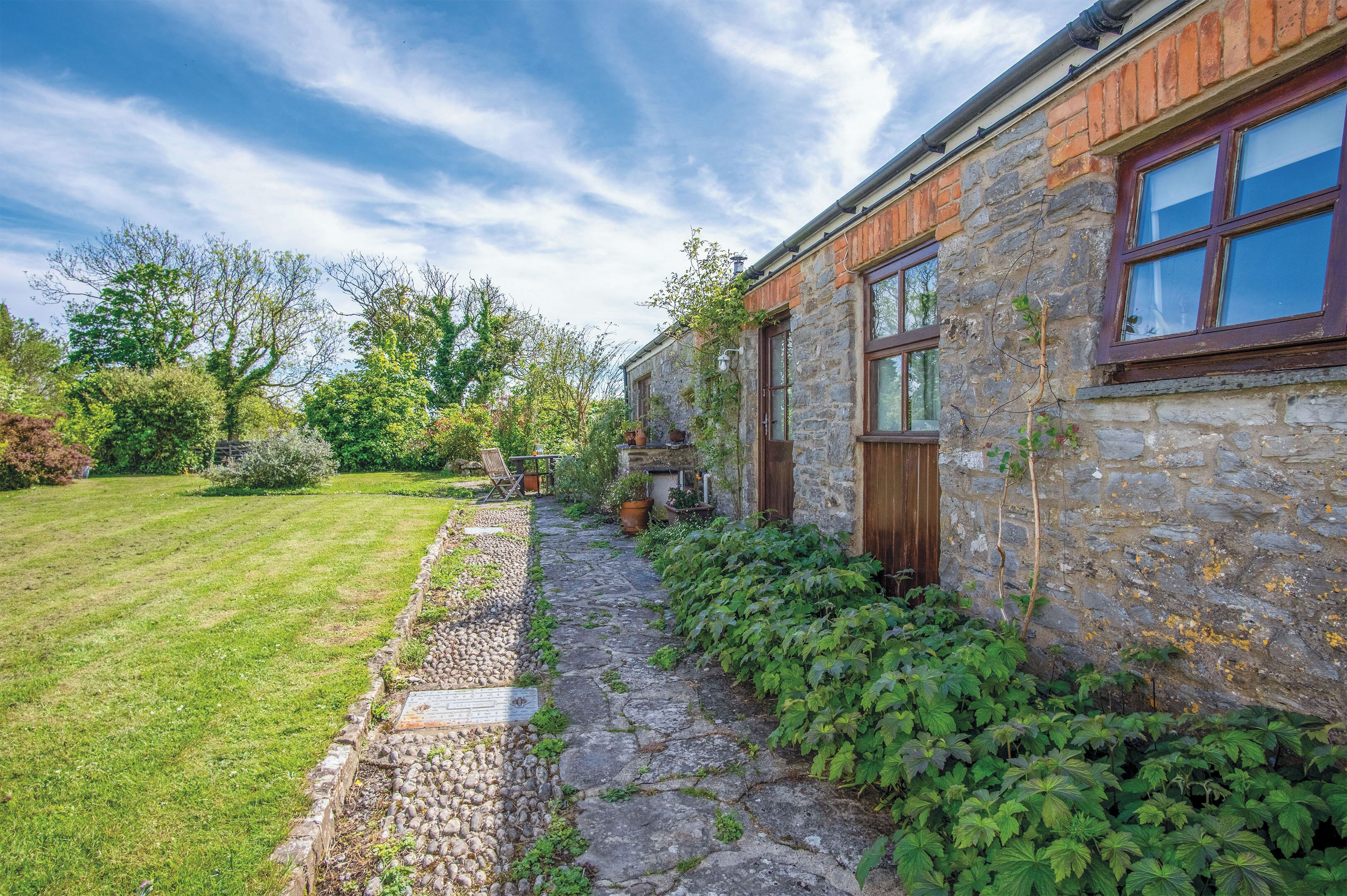Un bâtiment en pierre avec une porte et des fenêtres en bois, un chemin en pierre et une cour couverte d’herbe.