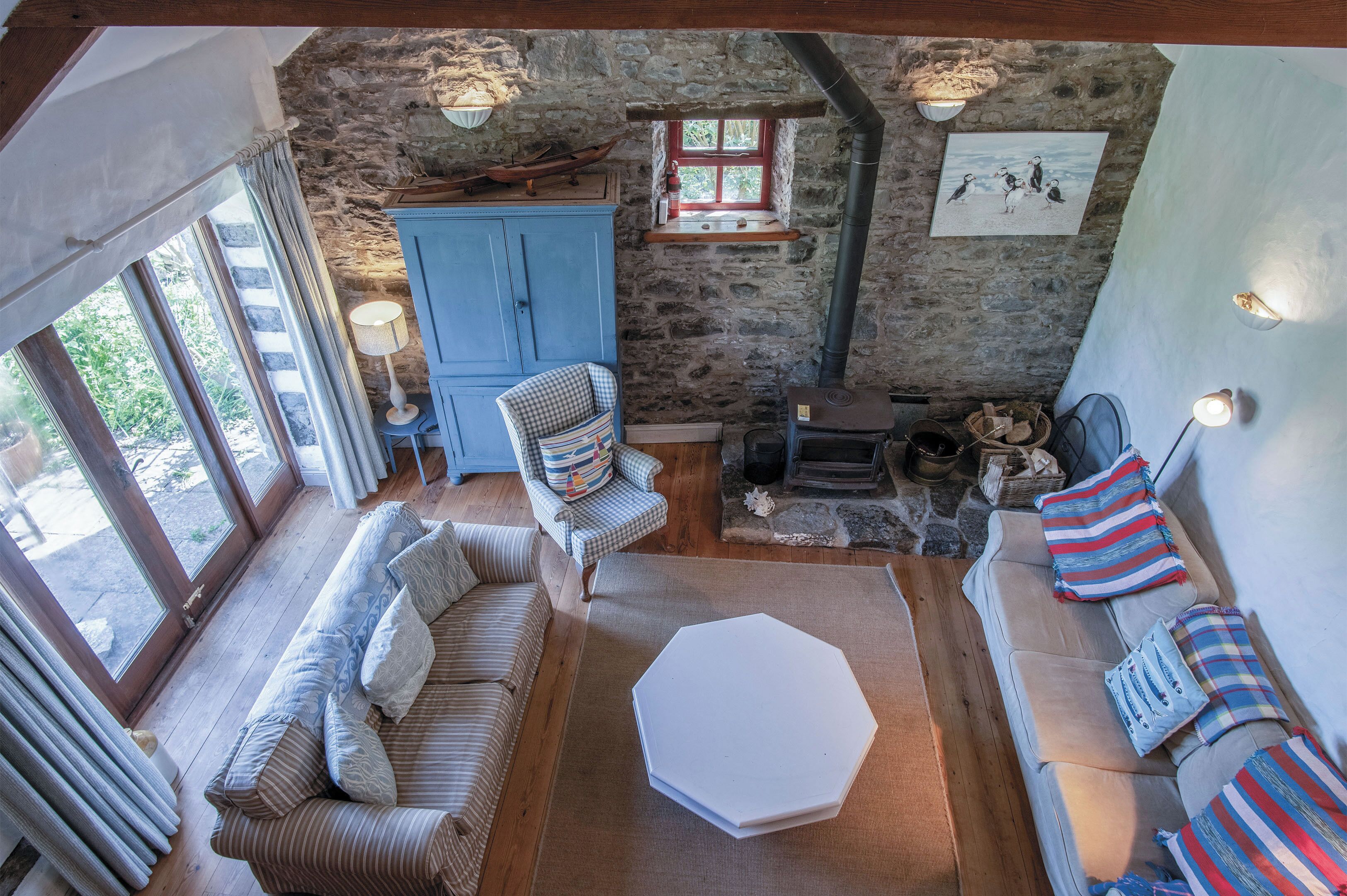 A cozy room with a stone wall, wooden furniture, a fireplace, and a window overlooking greenery.