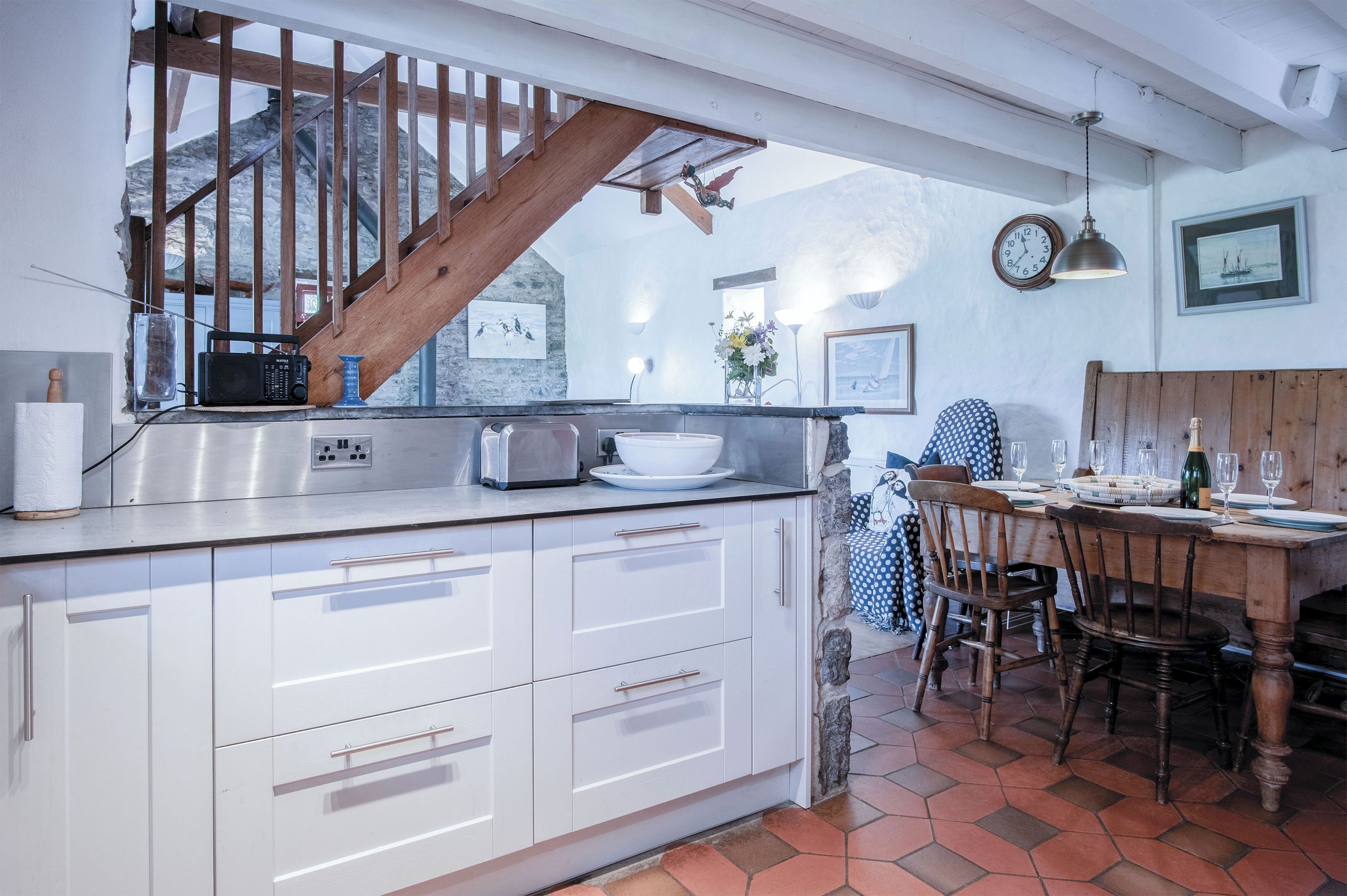 A kitchen with white cabinets, a wooden staircase, a dining table set for four, and a stone wall.