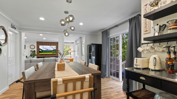 Un comedor moderno con una mesa de madera, sillas acolchadas, una lámpara de araña y una zona de cocina con estación de café.