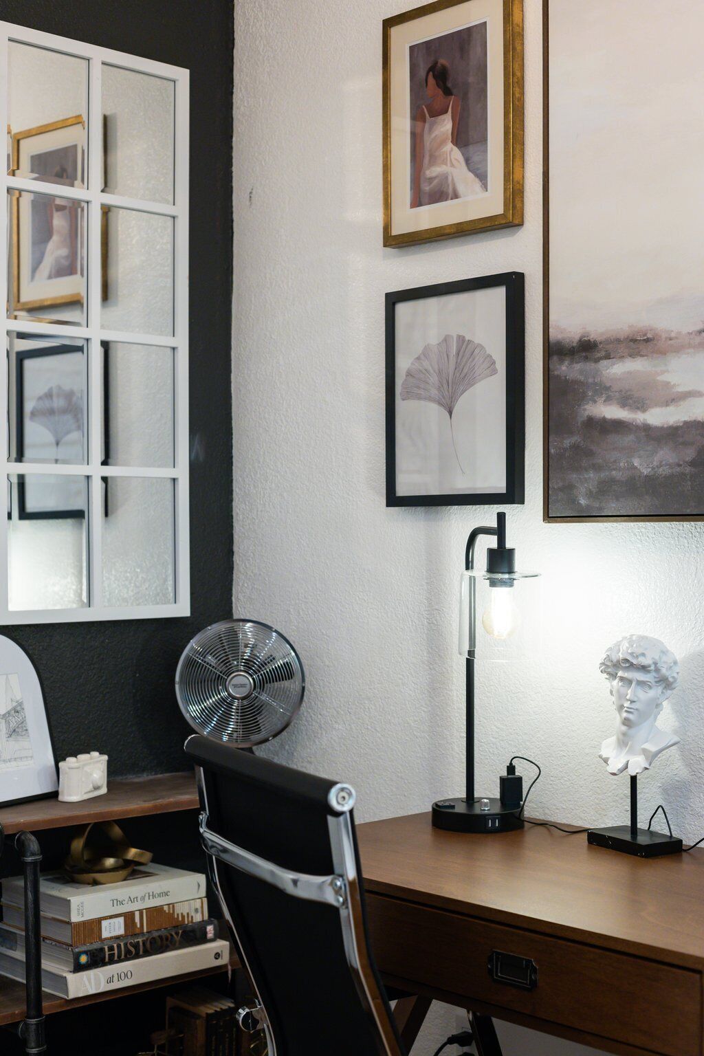 Una sala de estudio con un escritorio, una silla, libros, un ventilador, obras de arte enmarcadas y un busto en la pared.