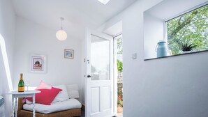 Um quarto claro com claraboia, porta branca que dá para o exterior, mesa pequena com garrafa e copo, e sofá de vime com almofadas.