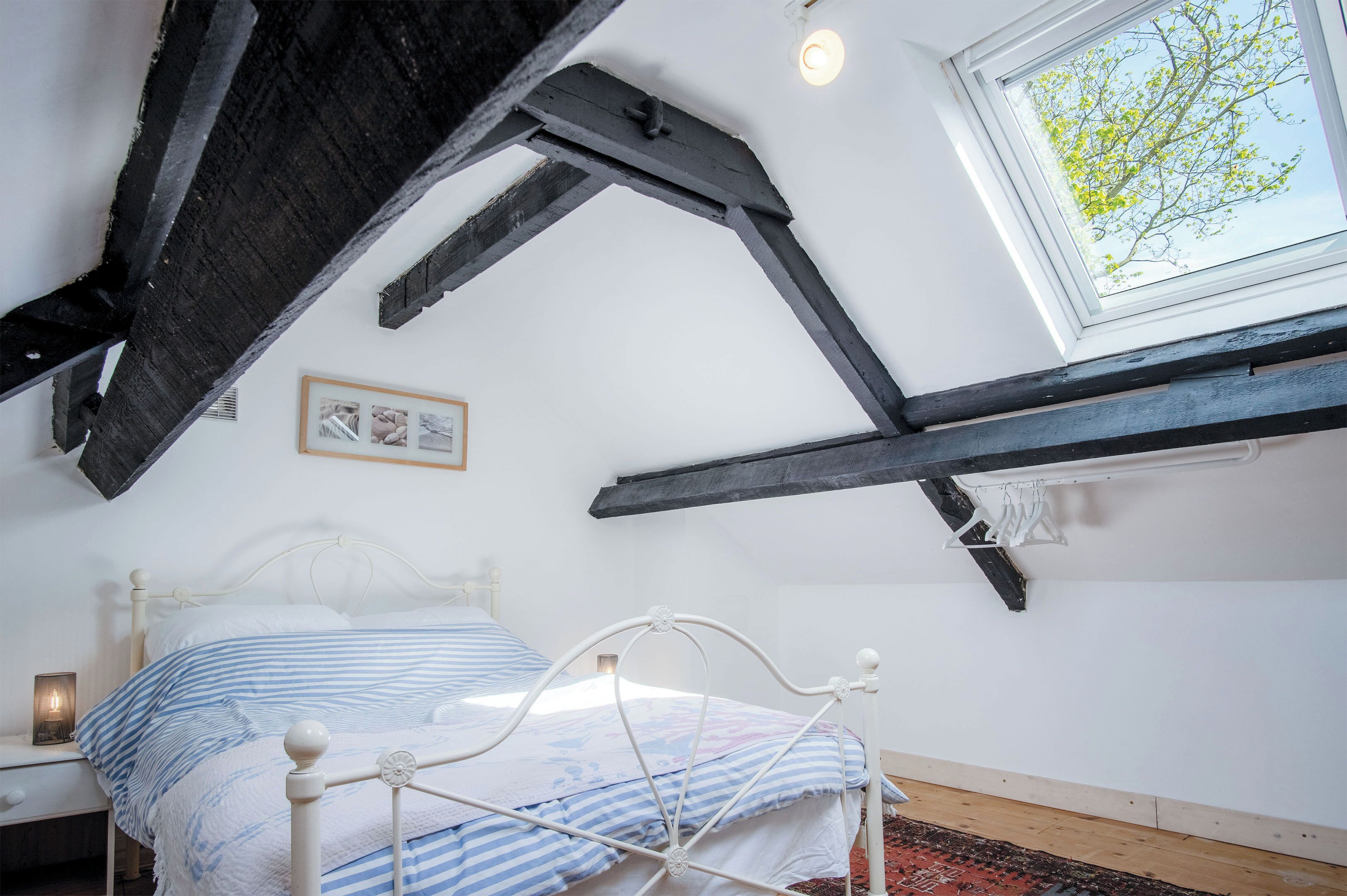 A bedroom with a sloped ceiling, a bed with a white metal frame, a window with a view of trees, and a framed picture on the wall.
