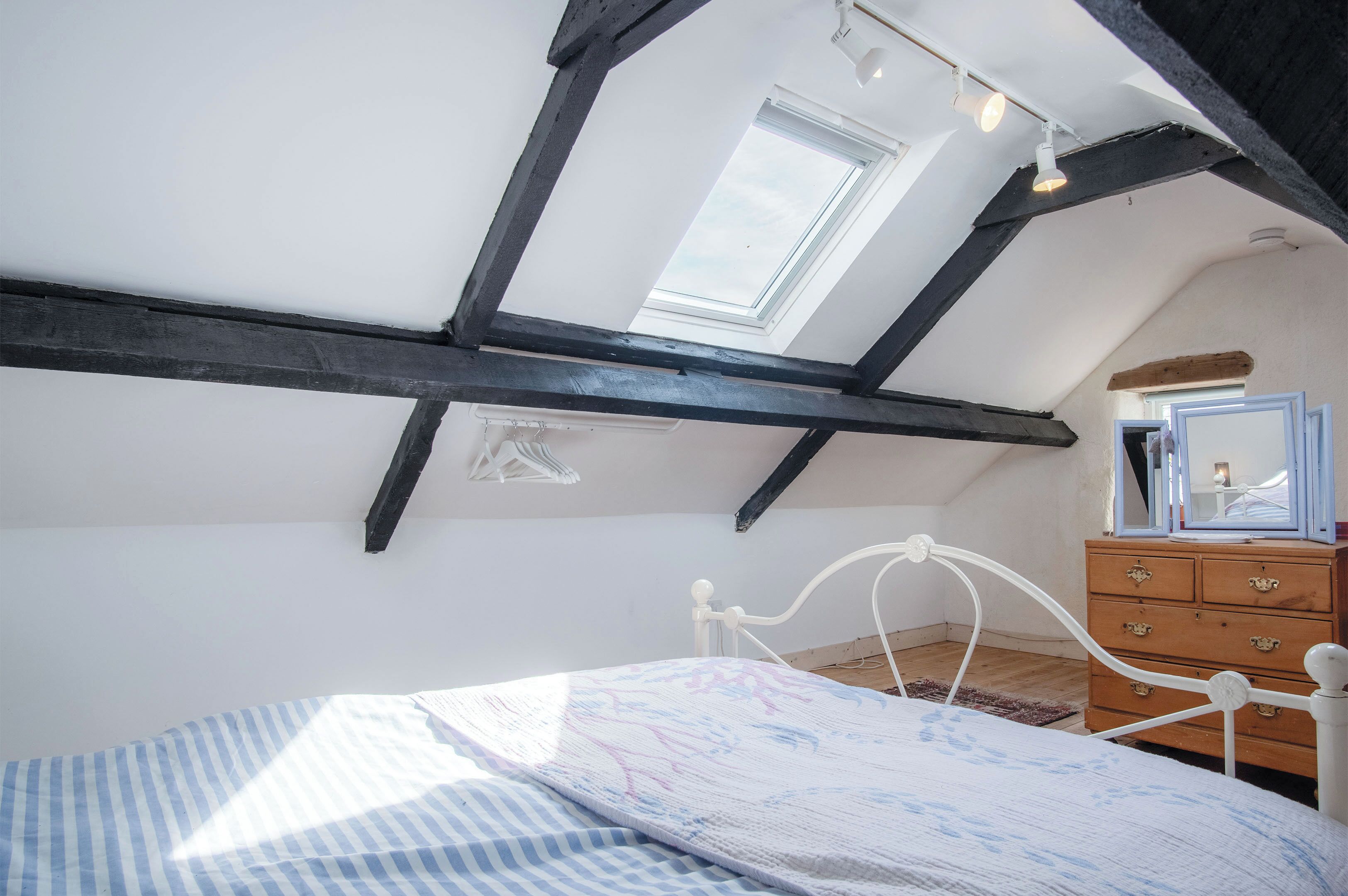 A bedroom with a sloped ceiling, a skylight, a bed with a striped bedspread, and a wooden dresser with a mirror.