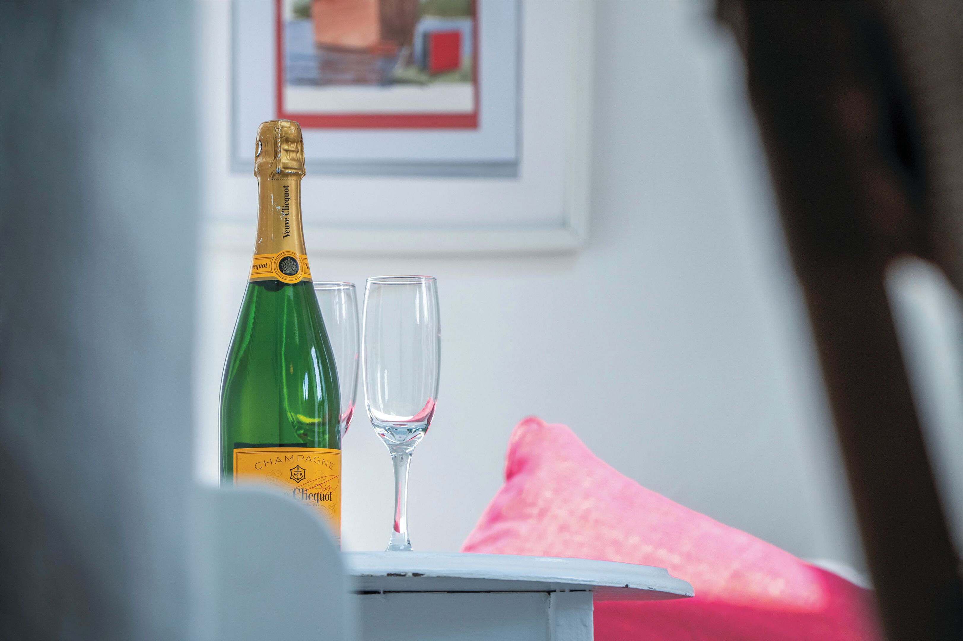 A bottle of champagne and two glasses on a table with a pink pillow in the background.