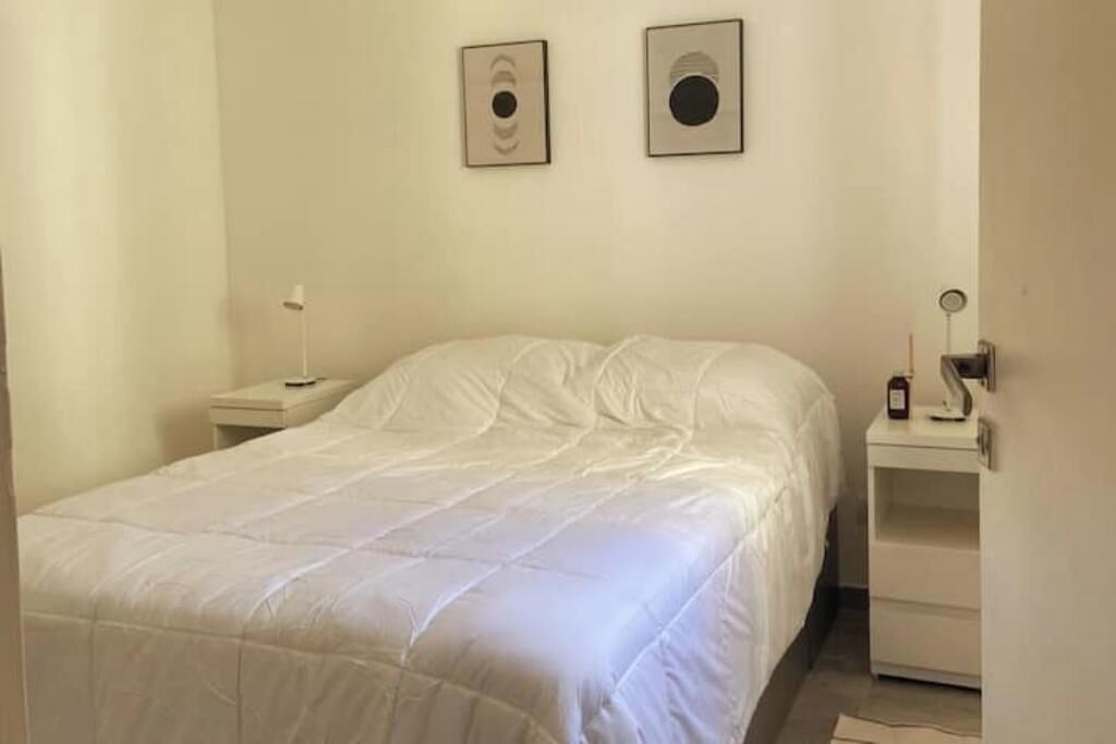 Uma cama bem arrumada em um quarto com esquema de cores branco, uma mesa de cabeceira com abajur e duas fotos emolduradas na parede.