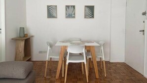 Un comedor con una mesa y sillas blancas, suelo de madera y tres obras de arte con motivos de hojas.