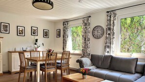 A modern living room with a dining area, featuring a wooden table, chairs, a sofa, and a coffee table. There are framed pictures on the wall, a clock, and large windows with curtains.