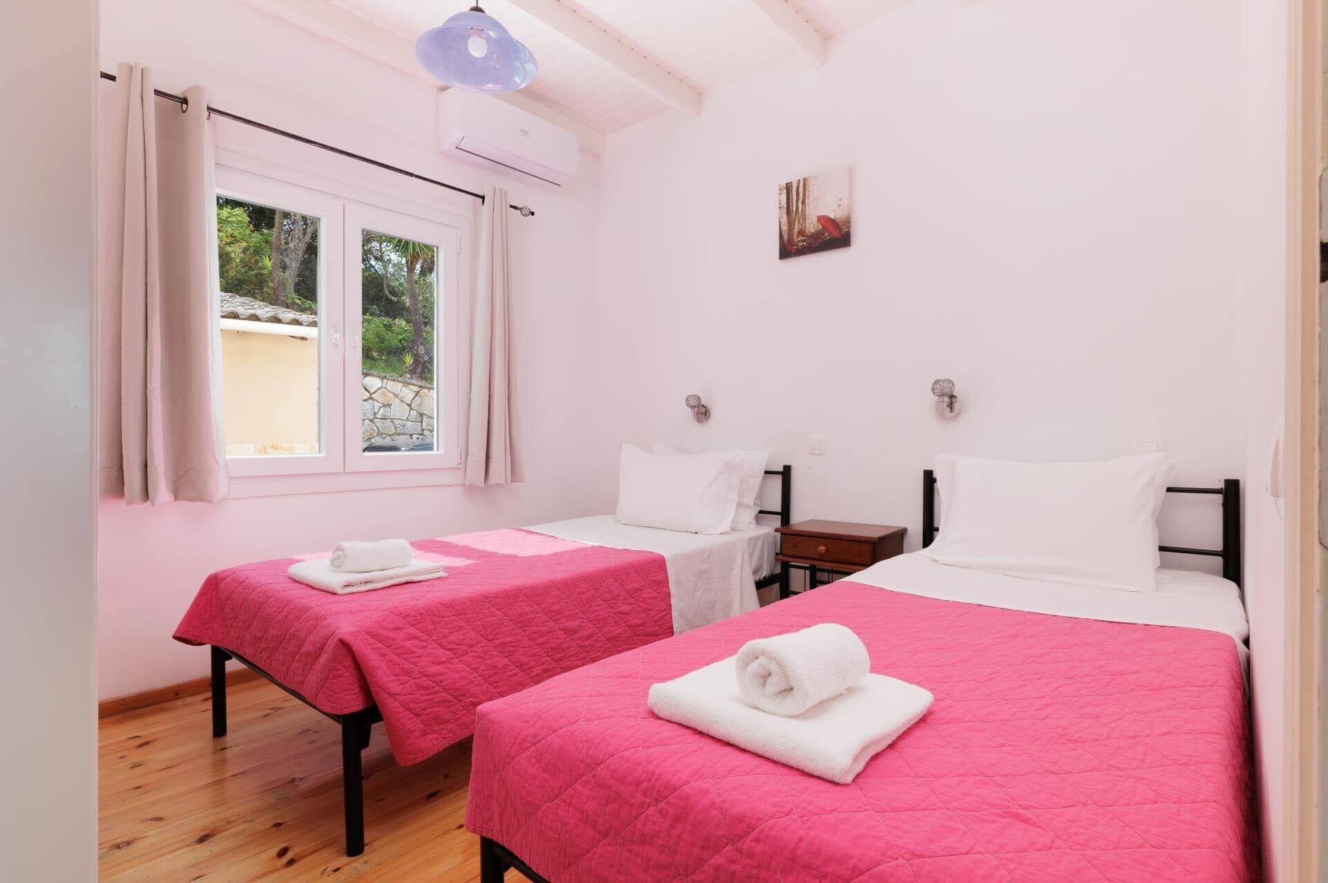 Une chambre avec deux lits, chacun recouvert d’une couette rose et agrémenté d’oreillers blancs, une fenêtre avec des rideaux et un luminaire fixé au mur.