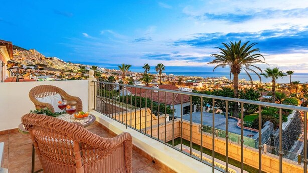 A balcony with wicker furniture, a table with fruit and a drink, and a view of a coastal town with palm trees and buildings. - Villa Costa Adeje by Villa Plus (Adeje)