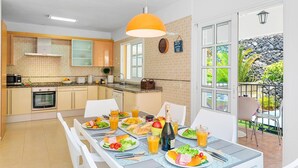 A modern kitchen with a dining area, featuring a large window, a dining table set with food, and a view of a garden. - Villa Costa Adeje by Villa Plus (Adeje)