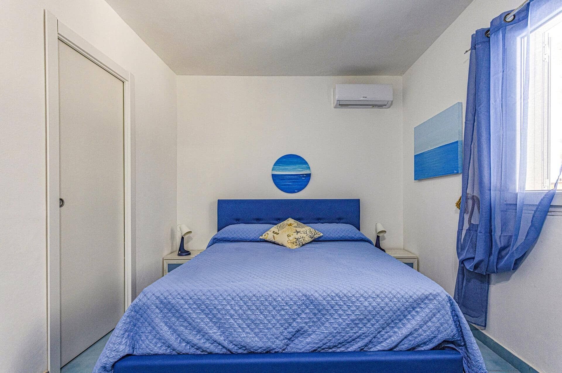 A bedroom with a single bed, a nightstand, a wall-mounted air conditioner, and a window with blue curtains.