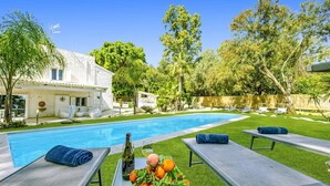 Ein Poolbereich mit Liegestühlen, einem Tisch mit Früchten und einer Flasche, sowie ein Haus im Hintergrund.