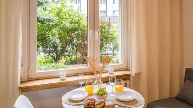 A dining area with a table set for two, a window with curtains, and a sofa in the background. - Kraków Czyżyny - Terrace & Workspace - by Rentujemy (Kraków)