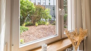 A room with a window overlooking a garden and a building. There is a table with a vase of dried flowers and a glass of orange juice. - Kraków Czyżyny - Terrace & Workspace - by Rentujemy (Kraków)