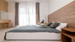 A modern bedroom with a large bed, wooden headboard, a bedside table with a lamp, and a view of the outdoors through sheer curtains. - Witolda Riverside Apartments - Balcony, City View - by Rentujemy (Wroclaw)