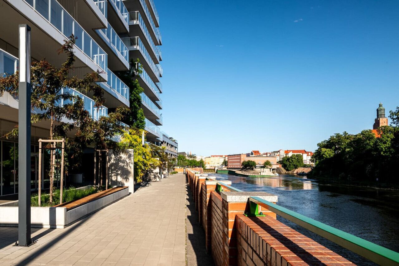 A modern building with a river view, a brick pathway, and a bridge.