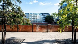 A paved walkway with a brick wall and trees on one side, leading to a modern building with multiple windows and balconies. - Witolda Riverside Apartments - Balcony, City View - by Rentujemy (Wroclaw)
