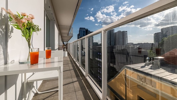 A balcony with a glass railing, a table with a vase of flowers and two glasses of orange juice, and a cityscape view. - Witolda Riverside Apartments - Balcony, City View - by Rentujemy (Wroclaw)