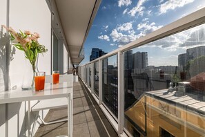 A balcony with a glass railing, a table with a vase of flowers and two glasses of orange juice, and a cityscape view. - Witolda Riverside Apartments - Balcony, City View - by Rentujemy (Wroclaw)