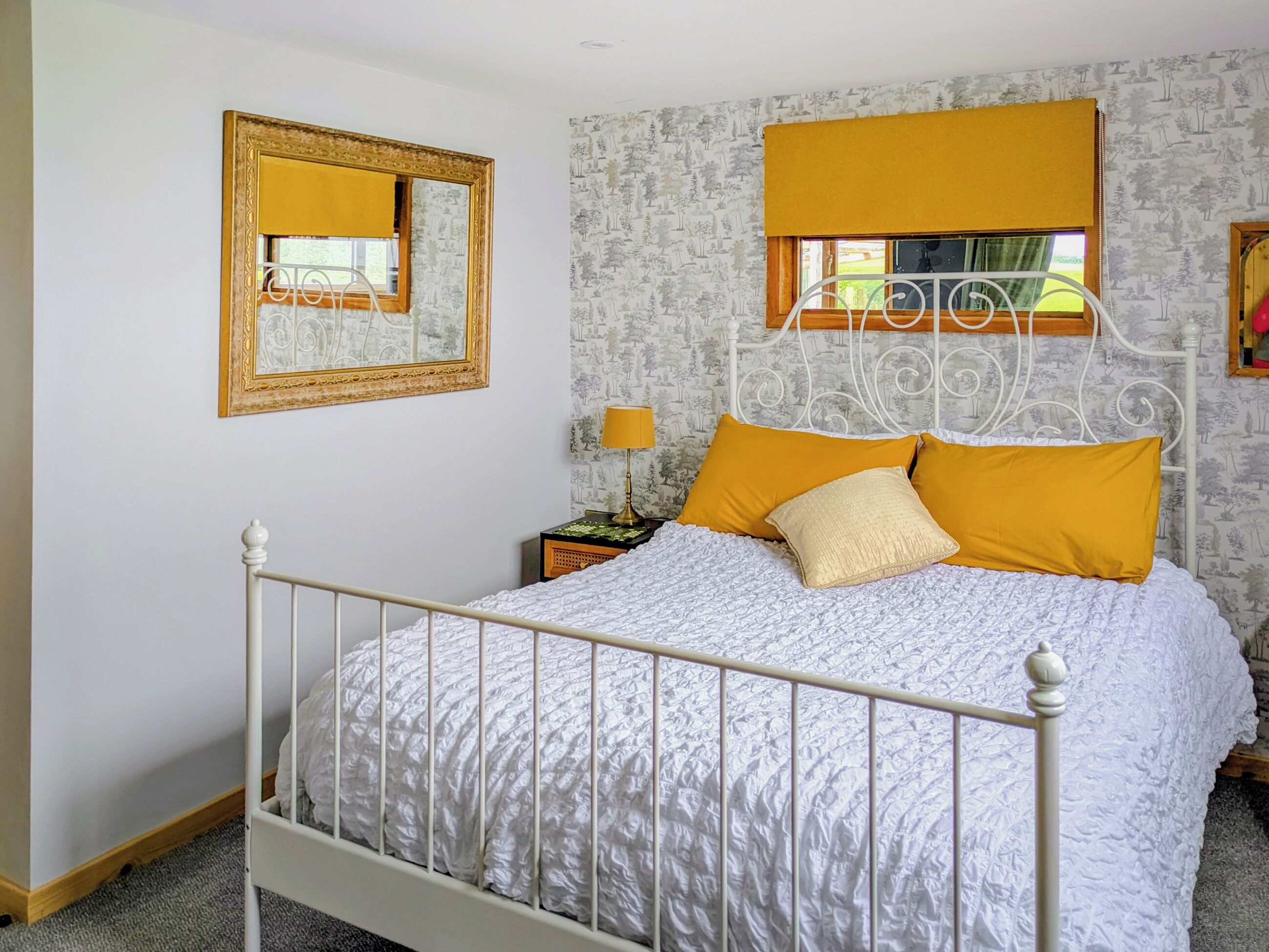 Camera da letto con un letto a letto in metallo bianco, una parete decorata, uno specchio e una finestra con persiana gialla.