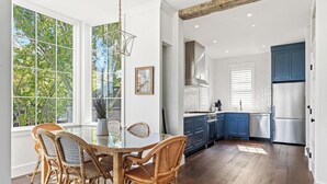Un comedor con una mesa de vidrio y sillas de mimbre, una cocina con armarios azules y electrodomésticos de acero inoxidable, y vistas a árboles a través de amplias ventanas.