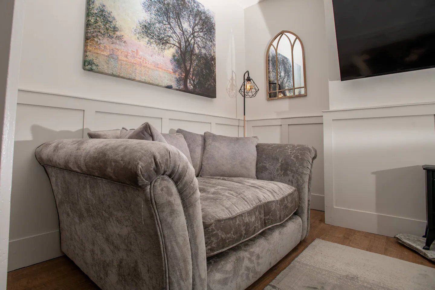 A living room with a grey sofa, a painting of a tree, a floor lamp, and a TV mounted on the wall.