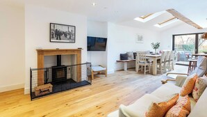 A spacious living room with a fireplace, wooden flooring, a dining area with a table and chairs, and a large window providing natural light.