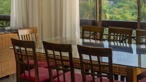 Un comedor con mesa de vidrio y sillas de madera con cojines rojos, ventanas amplias con cortinas, y lámparas colgantes.