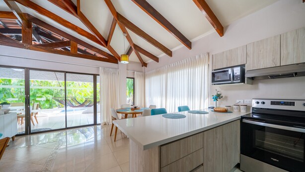 A modern kitchen with a large island, built-in microwave, and oven. There is a dining area with a table and chairs, and a view of greenery outside. - Villas Sol 21 Serenity Place at Playa Hermosa (Playa Hermosa)
