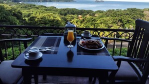 Ein Tisch mit Orangensaft, Kaffee und Essen auf einem Balkon mit Blick auf eine üppige Landschaft und das Meer.
