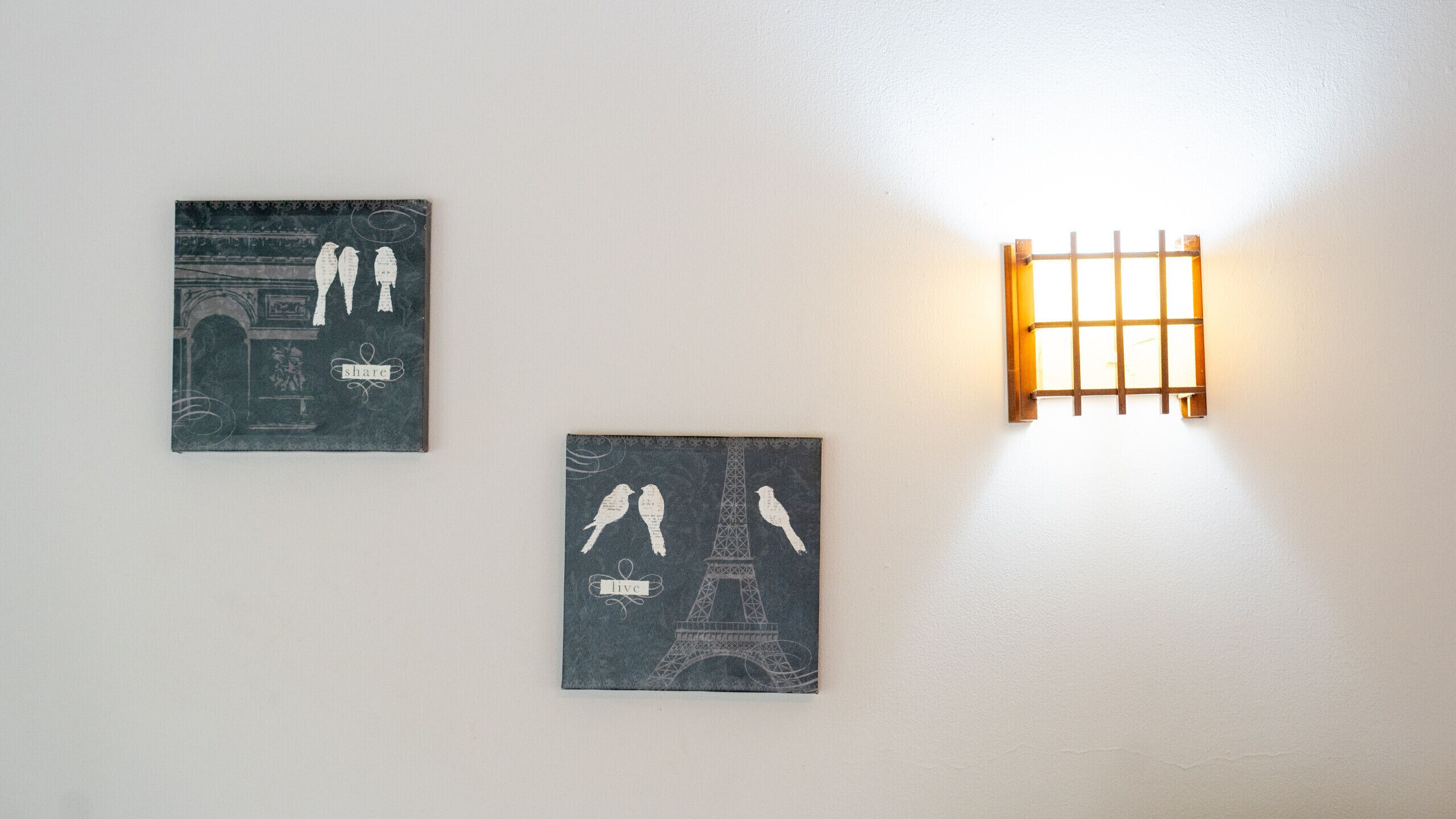 Two framed artworks on a wall, one depicting birds and the other the Eiffel Tower, with a light fixture above.