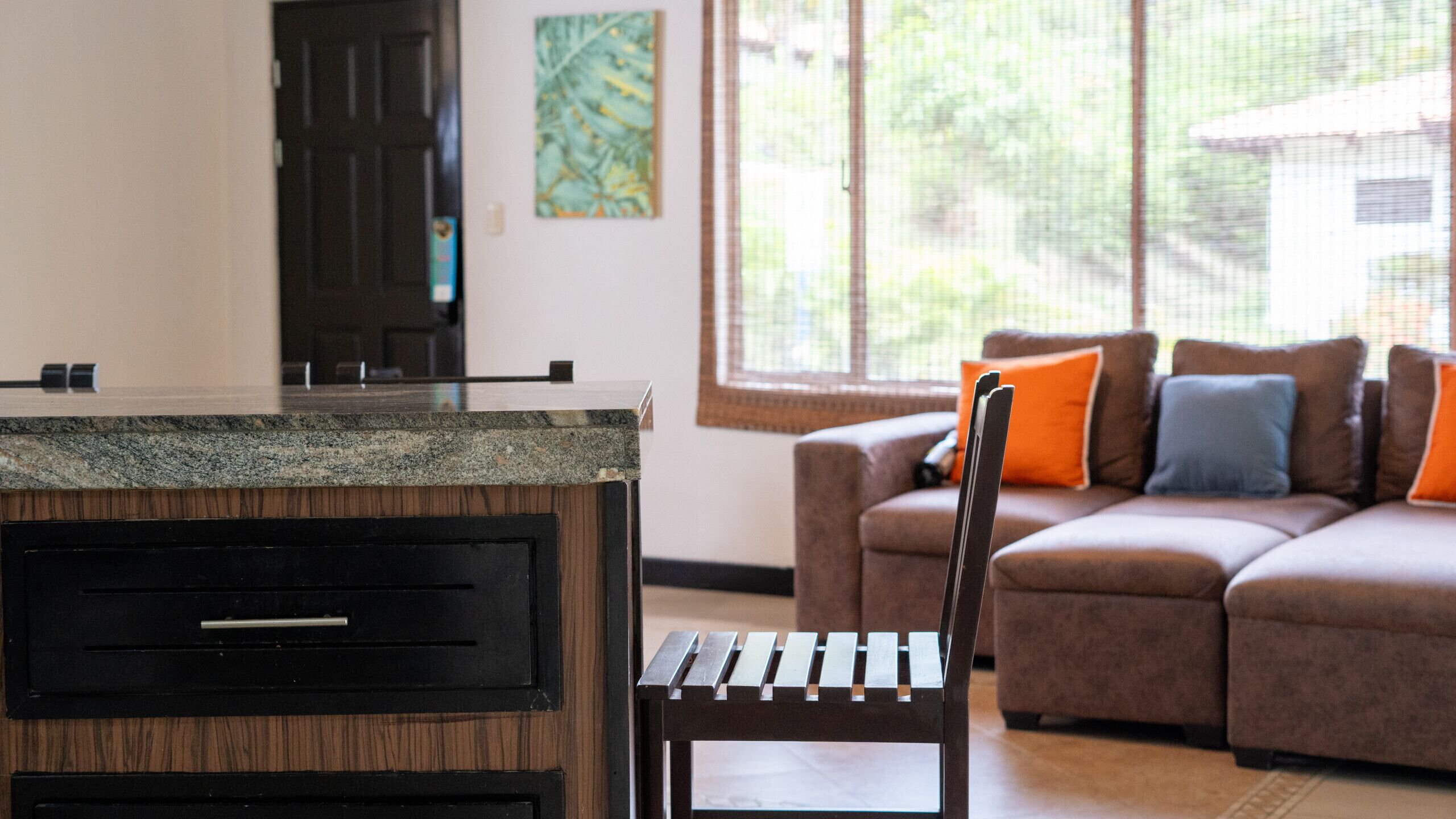 A kitchen with a granite countertop and a wooden cabinet. There is a dining chair with a metal frame and a cushion. A brown sofa with orange and blue cushions is in the background.
