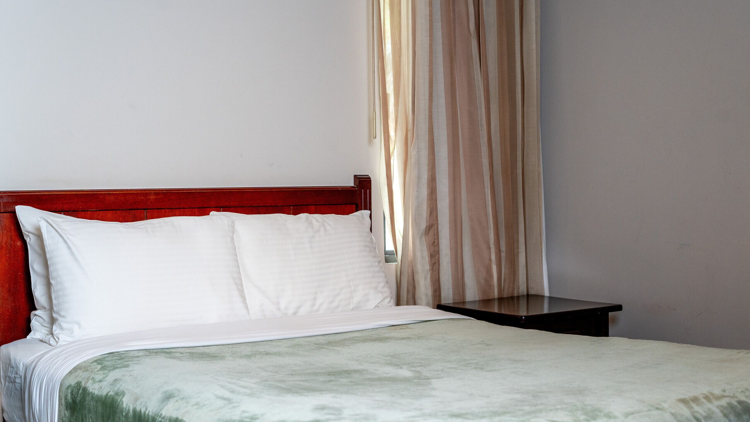 Una cama bien hecha con almohadas blancas y una manta verdosa, un cabecero de madera y una mesita de noche con una lámpara.