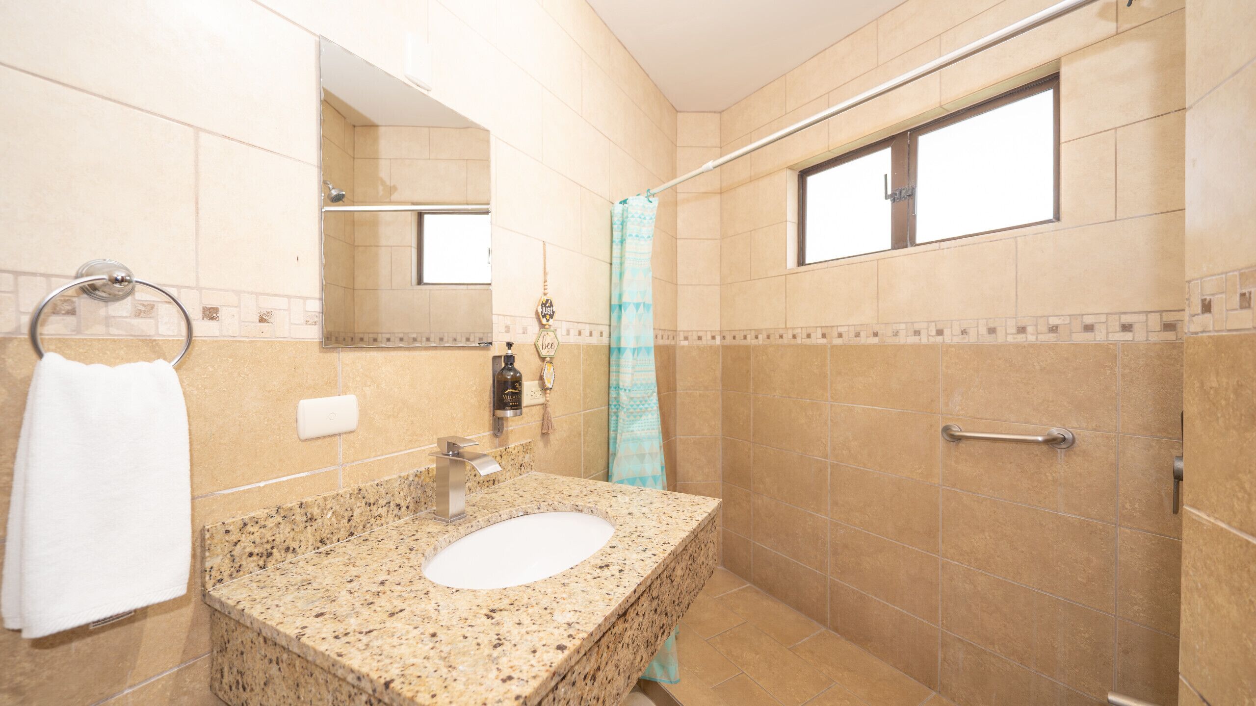 A bathroom with a granite sink, a mirror, a towel rack, a shower with a blue shower curtain, and a window.