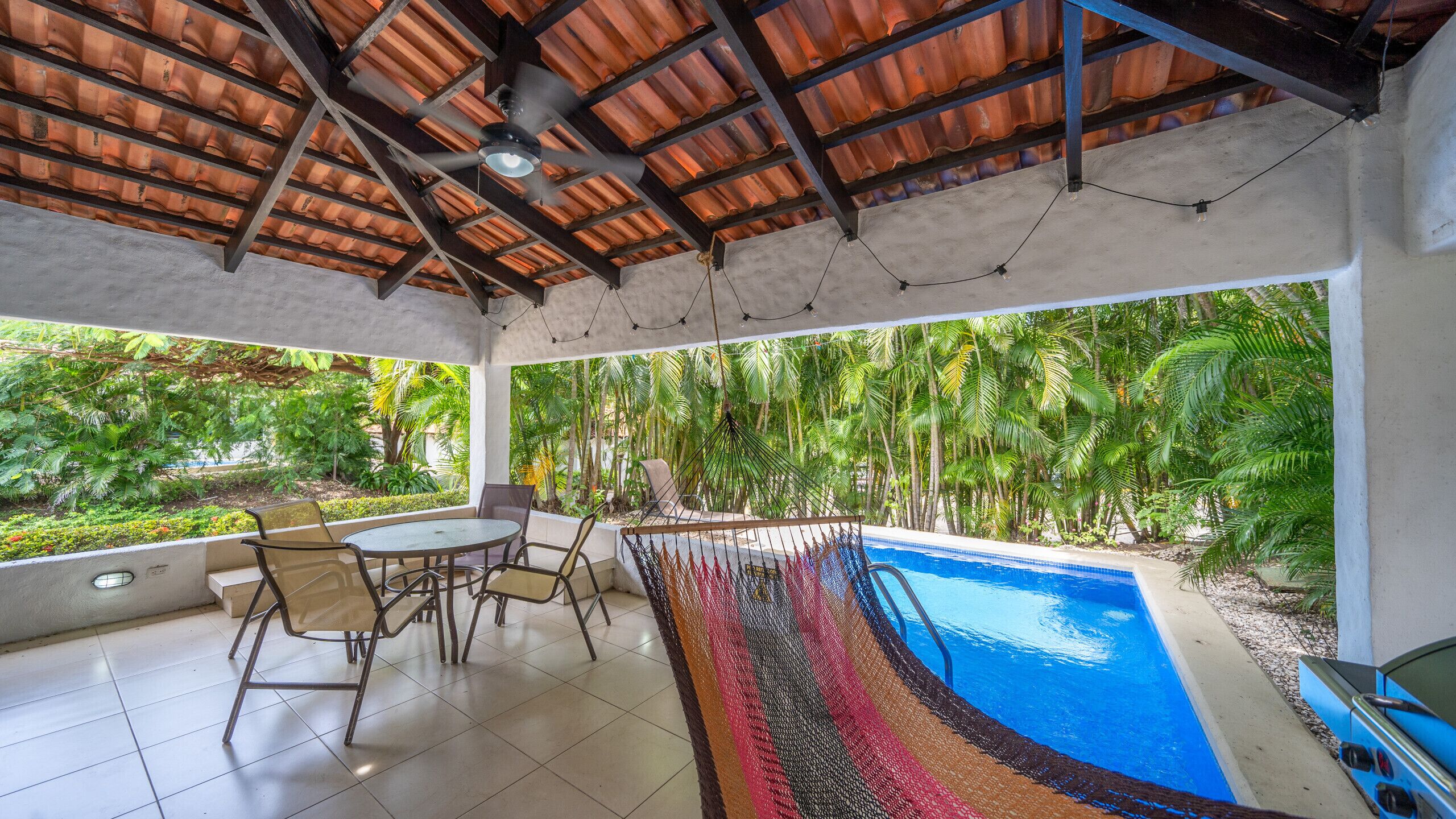 A covered outdoor area with a pool, seating, and a view of tropical vegetation.