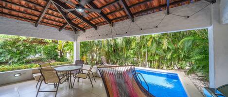 A covered outdoor area with a pool, seating, and a view of tropical vegetation.