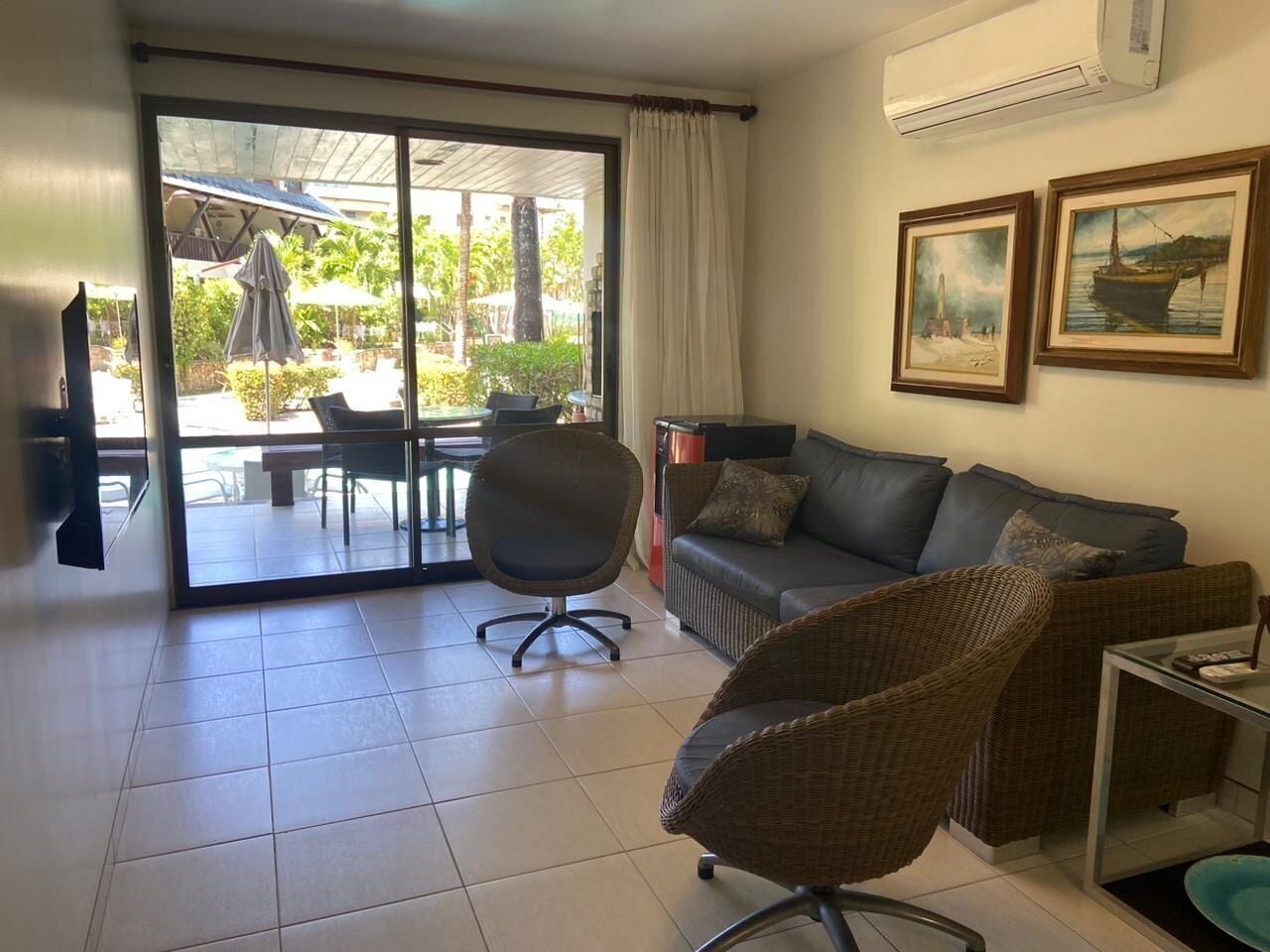 A living room with a sofa, armchair, and a glass coffee table. There is a sliding glass door leading to an outdoor patio area with a table and chairs.