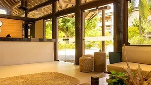 A modern interior with a wooden reception desk, a large window with a view of greenery, a seating area with a sofa and ottomans, and a wooden coffee table with a plant. - Condomínio Nannai Residence (Ipojuca)